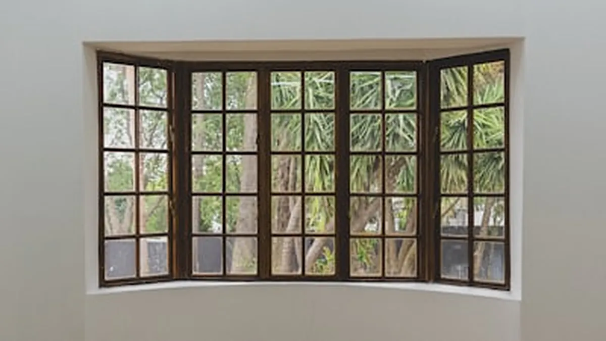Grande fenêtre en baie à cadre de bois, ouverte sur la verdure d’un jardin, dans une pièce aux murs clairs.